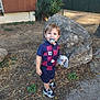 blue_clothing, building, child, daylight, ground, happy, leafy, maroon_clothing, number_10, outdoor, pacifier, person, plants, rock, shirt, shorts, smiling, sneakers, soccer_outfit, toddler