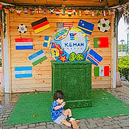 Muhammad is registered to the contest to win money with this photo: blue_shirt, child, daylight, decorations, event, festival, flags, green_carpet, holiday, international_flags, kemah, outdoor, pavement, person, playful, sculpture, sitting, soccer_ball, texas, wooden_wall