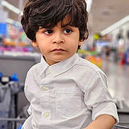 Muhammad is registered to the contest to win money with this photo: child, boy, shopping_cart, white_shirt, curly_hair, portrait, indoor, store, aisle, hand, expression, young, casual_clothing, retail, gaze, people, focus, lifestyle, cute, person