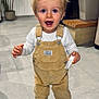 toddler, child, indoor, overalls, blonde_hair, blue_eyes, standing, floor, tiles, shoes, long_sleeve, holding, object, expression, person, home, plant, fireplace, background, curious