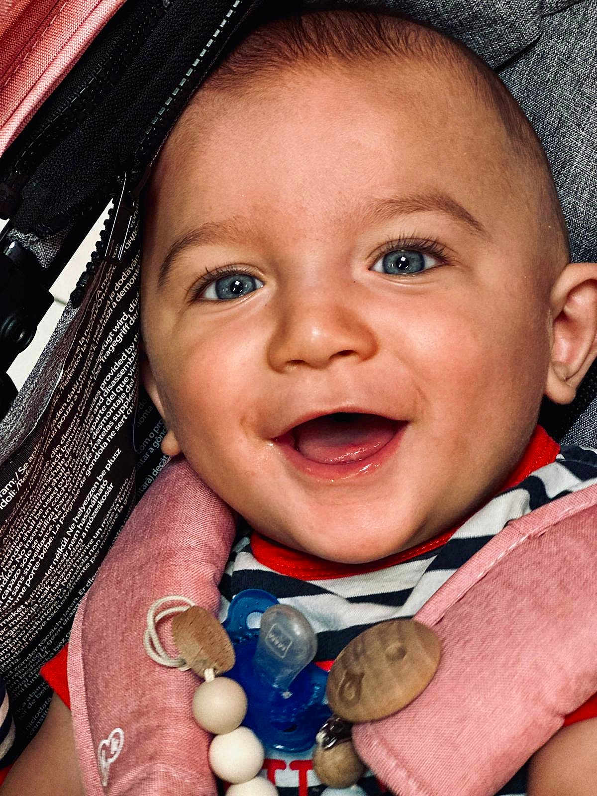 Julio participe au concours pour gagner de l'argent avec cette photo : baby, smiling, blue_eyes, pacifier, harness, striped_clothing, close_up, happy, infant, cute, child, face, mouth_open, pink, wooden_beads, cheerful, portrait, person, seat, accessory