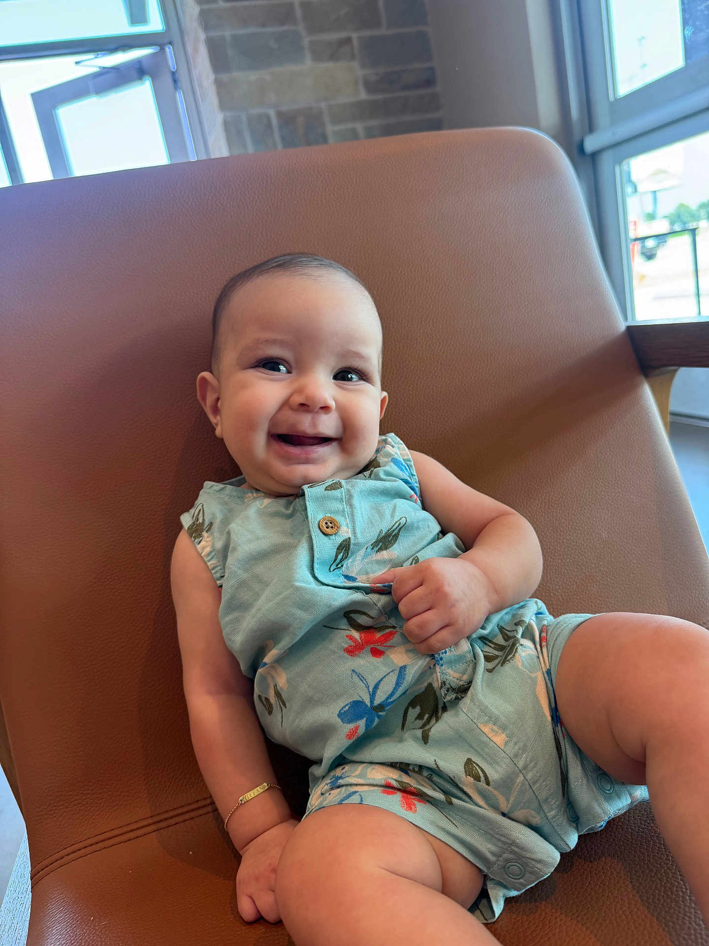 Stefano is registered to the contest to win money with this photo: baby, smiling, child, romper, floral_pattern, chair, indoor, natural_light, happy, cute, skin, arm, leg, bracelet, window, modern_furniture, casual, portrait, infant, relaxed