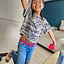 child, girl, smiling, glasses, braids, jeans, camouflage_shirt, pink_belt, pop_it_toy, indoor, wooden_floor, happy, playful, casual_clothing, footwear, earrings, young_person, portrait, dancing, fun