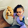 casual_clothing, child, cozy, curly_hair, dog, gray_bed, hand_sign, happy, indoor, light_wall, outlet, pet, playful, portrait, purple_vest, reflection, relaxed, smiling, sunglasses, wooden_floor