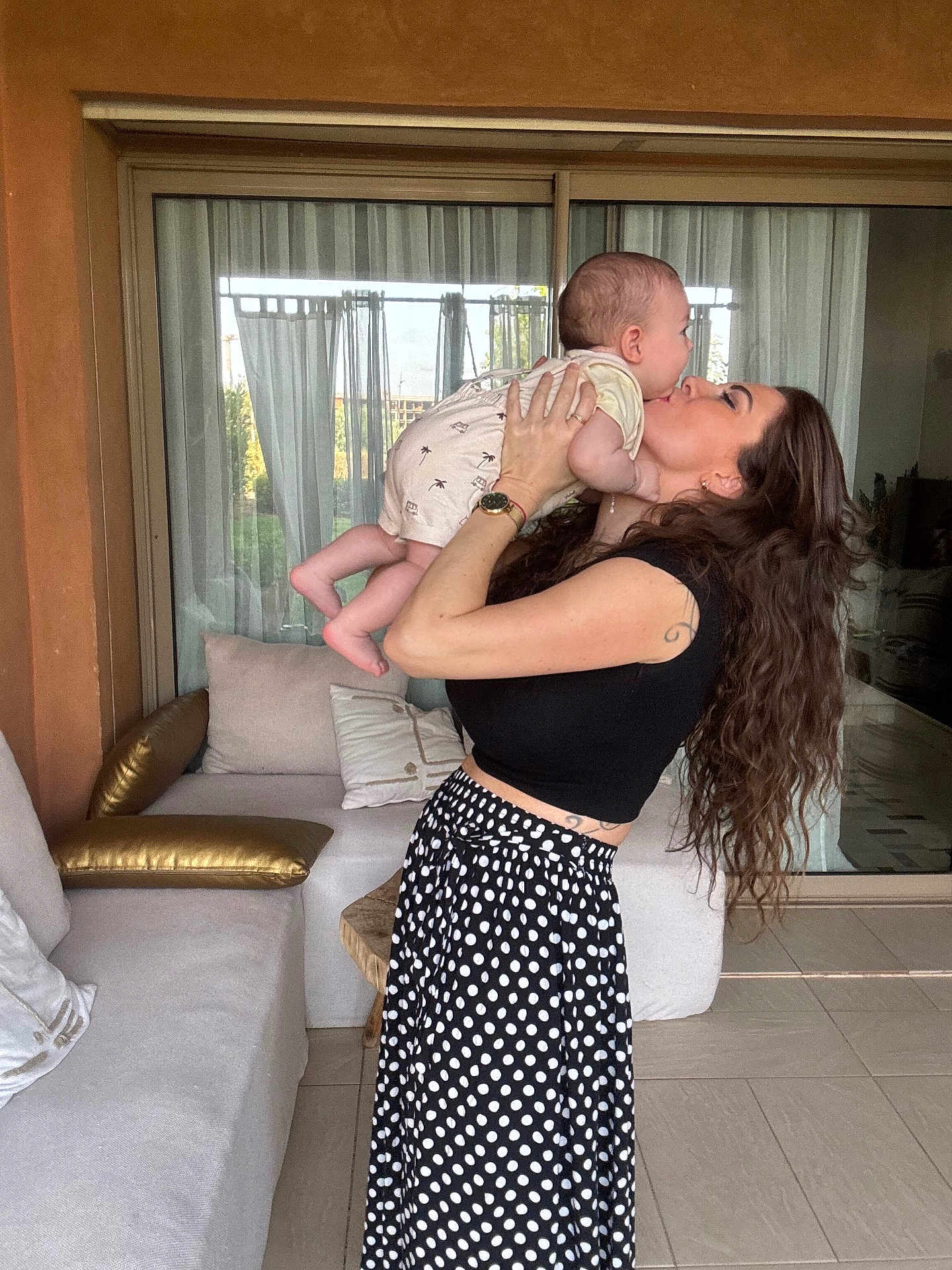 Naël participe au concours pour gagner de l'argent avec cette photo : affection, baby, casual_clothing, curtain, floor, happy, holding, indoor, kiss, long_hair, love, pillow, polka_dot, skirt, smile, sofa, tattoo, watch, window, woman