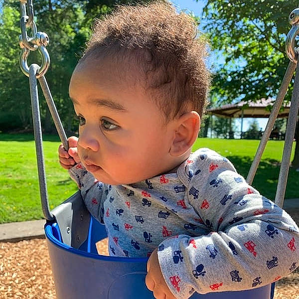 Orion is registered to the contest to win money with this photo: baby_toddler_clothing, chin, city, eye, fun, grass, green, happy, head, leisure, outdoor_play_equipment, person, plant, playground, public_space, recreation, sleeve, swing, toddler, tree
