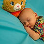 baby, sleeping, plush_toy, cat_toy, blue_background, colorful_shirt, floral_pattern, infant, peaceful, cute, soft_texture, child, portrait, resting, indoors, closeup, comfort, cuddly, toy, nap