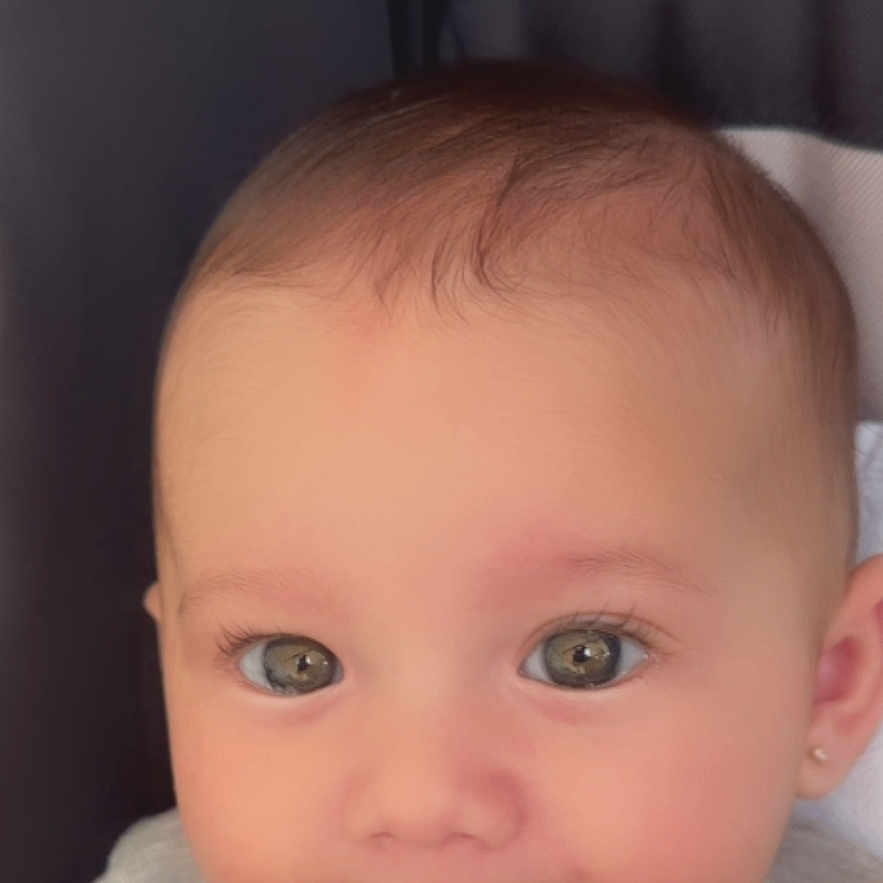 Griselda is registered to the contest to win money with this photo: baby, child, face, smile, drool, grey_clothing, cute, portrait, infant, head, eyes, earring, skin, sitting, indoors, young_child, cushion, soft_light, closeup, happy