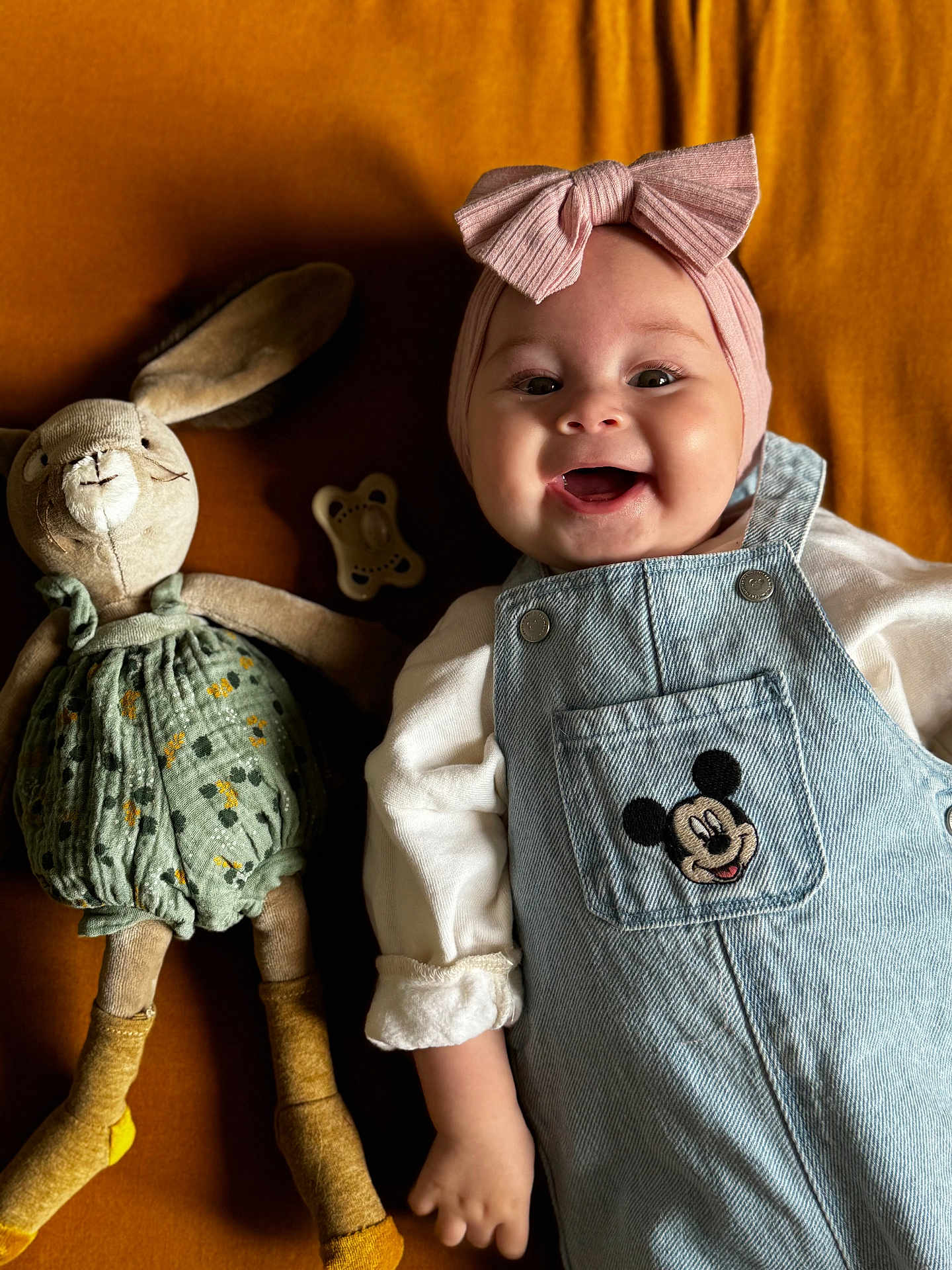 Haïvie participe au concours pour gagner de l'argent avec cette photo : baby, bow, bunny, child, clothing, cute, denim_overalls, happy, headband, indoor, infant, laying_down, mickey_mouse, mustard_background, pacifier, person, plush_toy, portrait, smiling, soft_toy