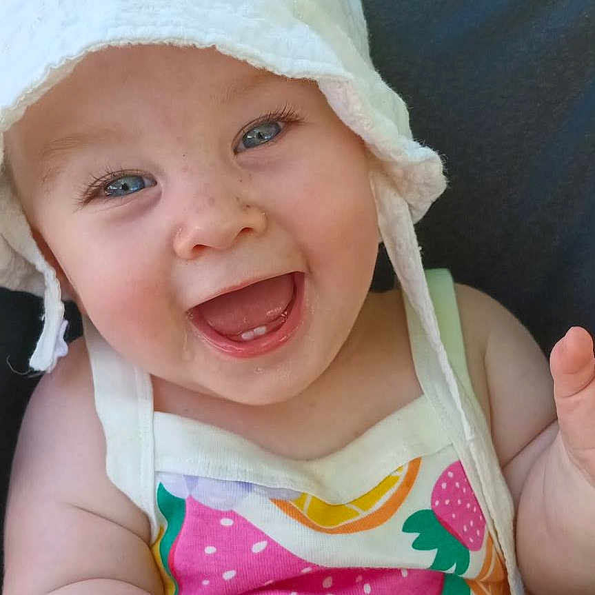 Novalie joined the competition — help win amazing prizes! baby, child, smiling, hat, white_hat, colorful_clothing, fruit_pattern, toy, outdoor, sunlight, happy, laughing, blue_eyes, cute, infant, person, summer, playful, sitting, closeup
