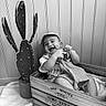 baby, smiling, cap, crate, wooden_crate, cactus, plant, indoor, blanket, cloth, child, happy, monochrome, black_and_white, floor, wall, pot, sitting, cute, portrait