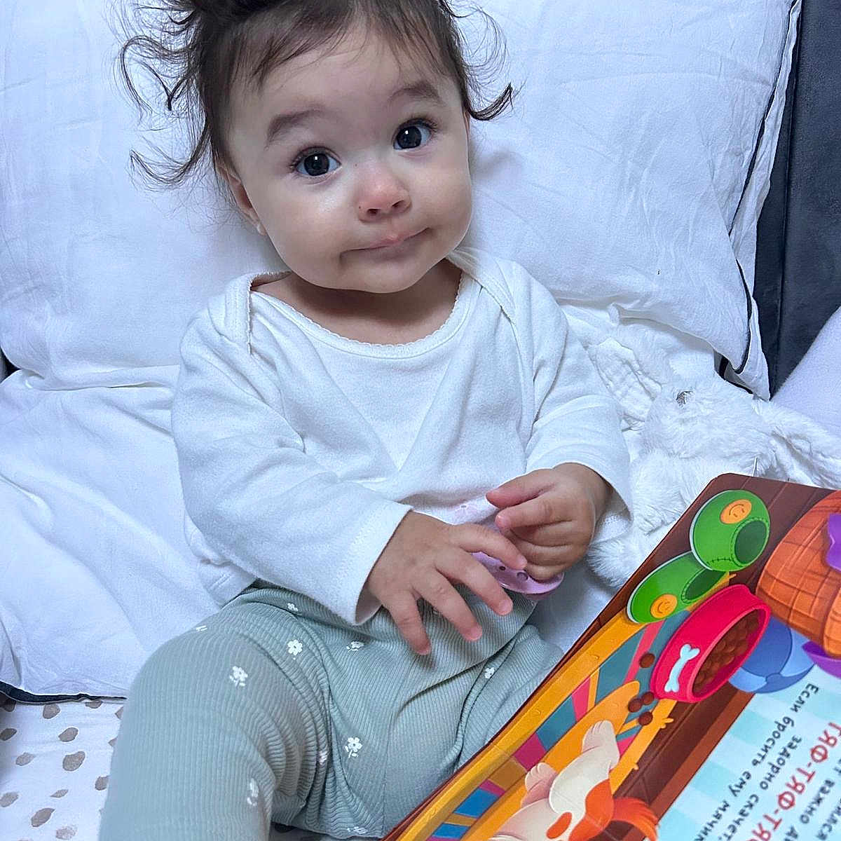Maya a rejoint le concours — aidez-le/la à gagner de superbes lots ! baby, bed, book, child, clothing, colorful, cute, expression, face, hair, indoor, patterned_pants, pillow, portrait, sitting, smiling, soft_toys, toddler, toy, white_shirt