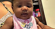 Loyal is registered to the contest to win money with this photo: adult, baby, child, curly_hair, dark_skin, eyes, face, floral_bib, hands, indoors, infant, jar, kitchen, lap, leopard_print, pigtails, pink_bib, portrait, shelf, sitting