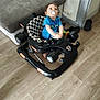 baby, child, walker, pacifier, blue_shirt, indoor, floor, couch, slippers, wooden_floor, toy, person, home, furniture, seat, black, white, checkered, looking_up, blur