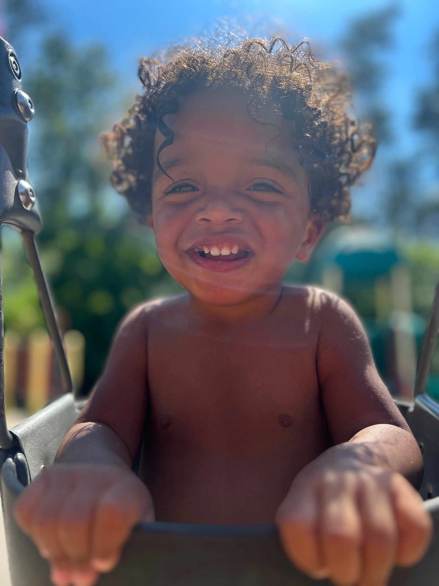 Orienn is registered to the contest to win money with this photo: child, toddler, smiling, curly_hair, swing, outdoor, playground, sunlight, happy, bare_chest, hands, daylight, blurred_background, nature, fun, joy, park, young_child, portrait, play