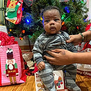 Nova joined the competition — help win amazing prizes! baby, child, christmas_tree, holiday, pajamas, striped_clothing, gift_bags, stocking, hands, floor, wooden_floor, indoor, decorations, colorful_lights, festive, person, face, curious, seated, toys