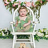 Willow is registered to the contest to win money with this photo: baby, child, green_dress, flower_crown, rocking_chair, flowers, pink_flowers, greenery, wooden_blocks, milestone, six_months, smiling, portrait, sitting, cute, indoor, decor, celebration, happy, infant