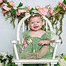 baby, child, smiling, green_dress, flower_crown, white_chair, flowers, pink_flowers, white_flowers, greenery, portrait, cute, happy, sitting, indoor, decorative, nature, flora, celebration, toddler