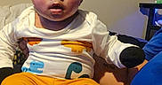 I'Zayvion joined the competition — help win amazing prizes! baby, infant, child, person, clothing, dinosaur_pattern, orange_pants, white_shirt, black_socks, indoor, window_air_conditioner, furniture, table, person_holding_baby, curly_hair, sitting, portrait, face, expression, home