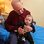 Haidyn joined the competition — help win amazing prizes! elderly_man, baby, indoor, holding, plaid_shirt, black_clothing, pacifier, bed, blue_bedspread, generational_bonding, seated, warm_lighting, casual_clothing, hands, face, person, portrait, home, comfort, family