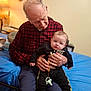 elderly_man, baby, indoor, holding, plaid_shirt, black_clothing, pacifier, bed, blue_bedspread, generational_bonding, seated, warm_lighting, casual_clothing, hands, face, person, portrait, home, comfort, family