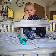 Colton is registered to the contest to win money with this photo: baby, hospital, hospital_bed, medical_equipment, monitor, infantino, baby_seat, child, bed, pajamas, person, indoor, medical_monitor, clock, hand, face, footwear, room, cable, furniture