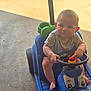Colton is registered to the contest to win money with this photo: baby, child, toy_car, blue, steering_wheel, outdoor, sunlight, concrete_floor, green, sitting, footwearless, cute, playtime, person, infant, toy, expression, vehicle, small, young