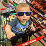 toddler, child, sunglasses, shopping_cart, store, snacks, shelf, clothing, blue_crocs, baby, person, retail, indoor, supermarket, toy, hand, floor, casual, cute, shopping