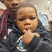 Jamari is registered to the contest to win money with this photo: toddler, child, adult, person, face, finger_in_mouth, hoodie, braids, indoor, room, wall_decoration, plastic_bag, shelf, clothing, portrait, casual, curious, black_hoodie, closeup, seated