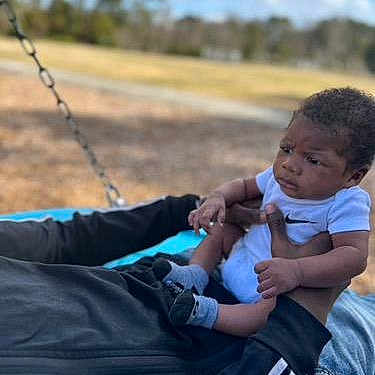 Jamari is registered to the contest to win money with this photo: toddler, child, outdoor, swing, sky, clouds, trees, park, grass, person, holding, baby, casual_clothing, daylight, expression, nature, recreation, playing, family, fun