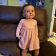 Taylor is registered to the contest to win money with this photo: toddler, child, pink_dress, brown_chair, living_room, standing, curious_expression, hair, face, barefoot, furniture, indoor, clothing, person, home, window, curtains, shelf, money, diaper
