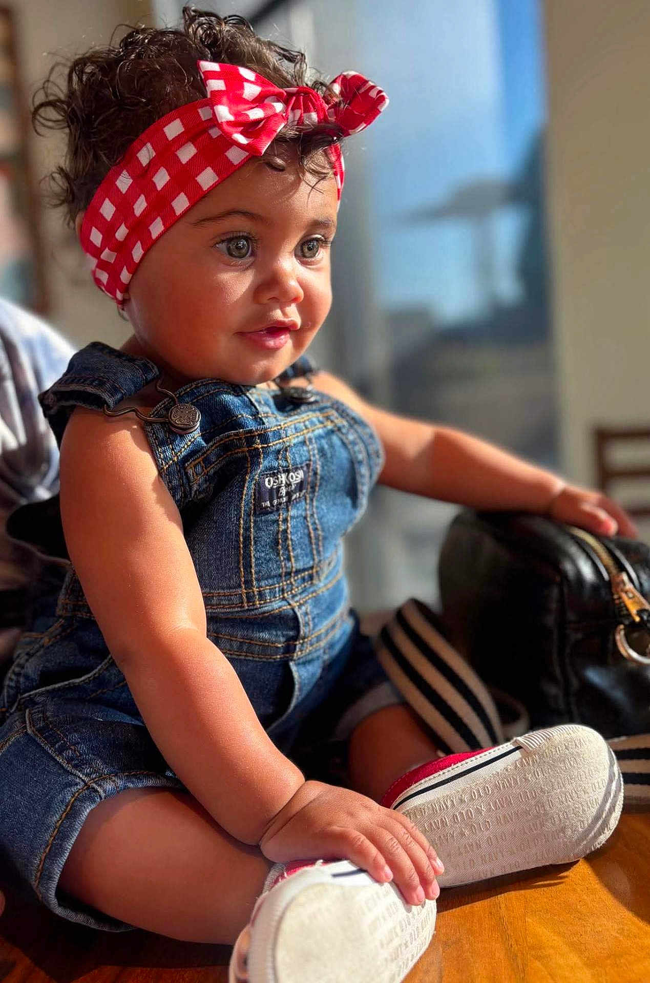 Emmy joined the competition — help win amazing prizes! child, toddler, denim_overalls, headband, red_checkered, curly_hair, indoor, sunlight, portrait, cute, smiling, baby_shoes, hand, face, sitting, person, young_child, casual_clothing, table, background_blur