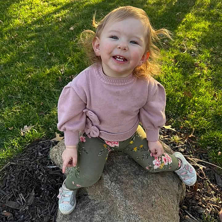 Caleigh joined the competition — help win amazing prizes! casual_clothing, child, daylight, flower_pattern, grass, happy, nature, outdoor, park, person, pink_sweater, playful, portrait, rock, shadow, smiling, sneakers, sunlight, toddler, young_child