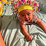 baby, infant, bed, blanket, pillow, bonnet, headband, pineapple_pattern, yellow, pink, blue, skin, tattoo, hand, face, expression, indoor, person, clothing, curious