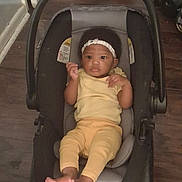 Miracle is registered to the contest to win money with this photo: baby, infant, car_seat, yellow_clothing, headband, indoors, wooden_floor, footwear_bare, child, person, seat, black, gray, curious, small, cute, looking, relaxed, casual, young