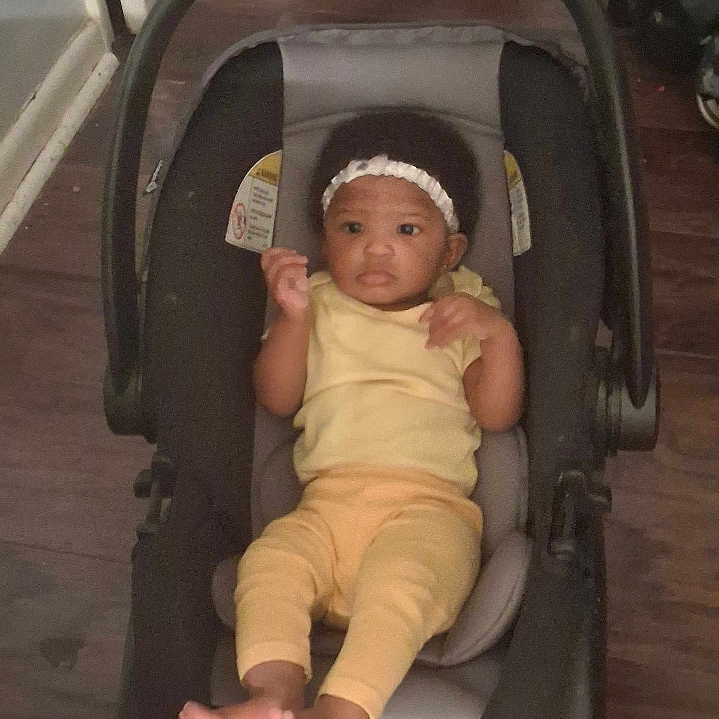 Miracle is registered to the contest to win money with this photo: baby, infant, car_seat, yellow_clothing, headband, indoors, wooden_floor, footwear_bare, child, person, seat, black, gray, curious, small, cute, looking, relaxed, casual, young
