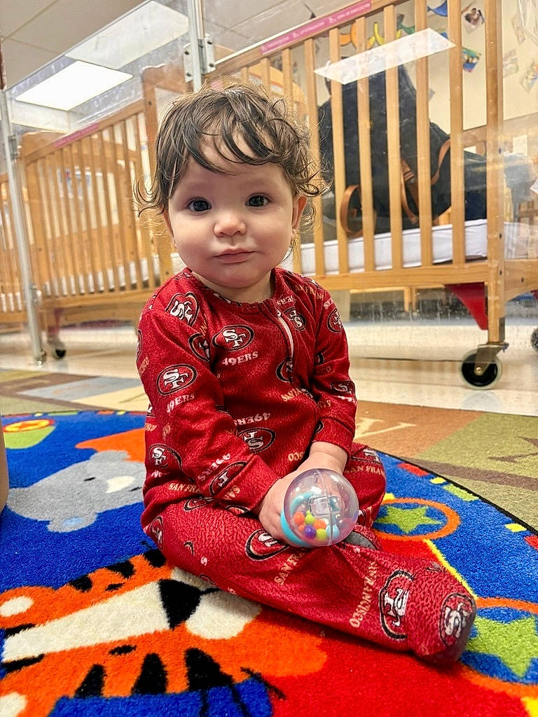 Allyana joined the competition — help win amazing prizes! baby, child, colorful_rug, crib, curly_hair, daycare, eyebrows, floor, indoor, nursery, pajamas, playroom, playtime, portrait, rattle, red_clothing, sitting, smile, toddler, toy_ball
