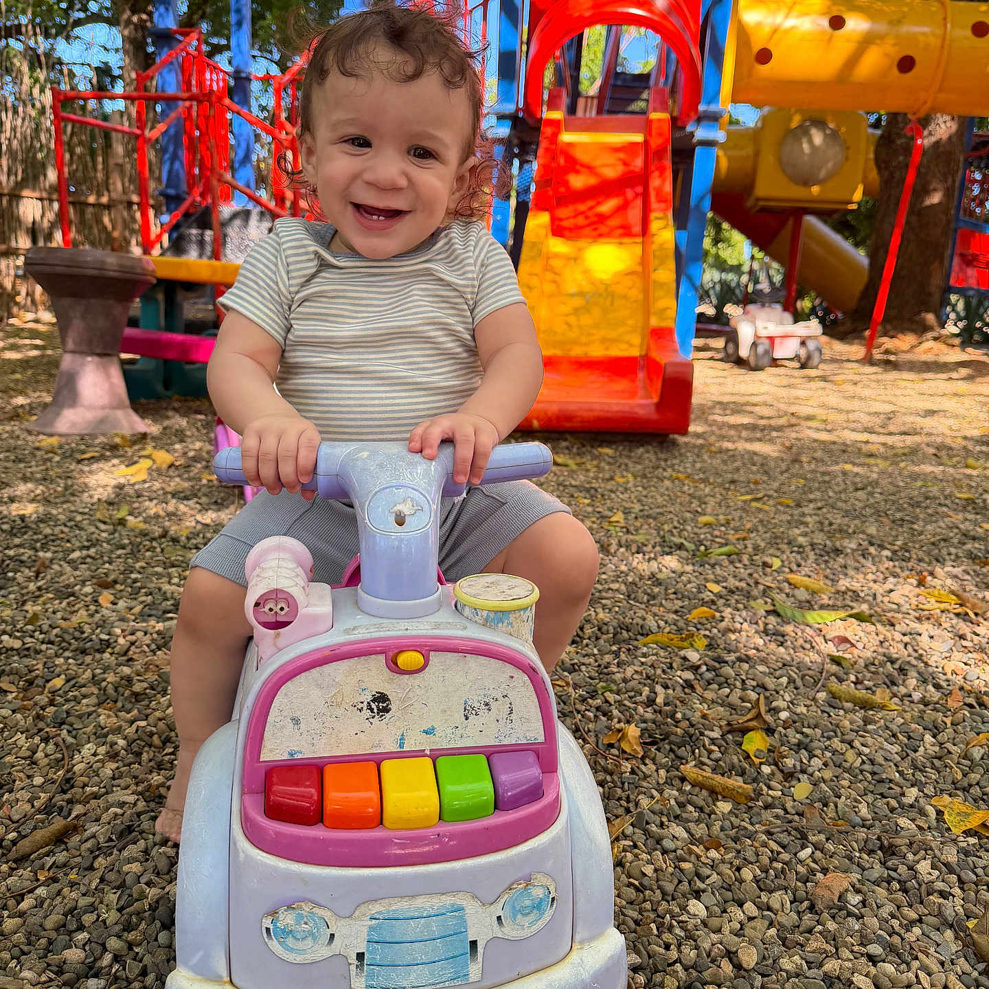 Oniel joined the competition — help win amazing prizes! child, climbing_structure, colorful, curly_hair, daylight, fun, gravel, happy, nature, outdoor, play, playground, shorts, slide, smiling, striped_shirt, sunlight, toddler, toy_car, trees