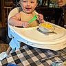 baby, high_chair, spoon, food, smile, messy, person, elderly, kitchen, tablecloth, whisk, bowl, ice_cube_tray, wooden_floor, glasses, facial_expression, feeding, indoor, happy, table