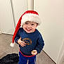 black_socks, blue_pants, blue_sweatshirt, casual, child, clothing, floor, happy, hat, holding_object, holiday, indoor, jacket, person, portrait, red_trim, santa_hat, smiling, standing, toddler