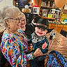 baby, child, elderly_woman, woman, cowboy_hat, bandana, smile, glasses, balloon, trophies, table, chair, bag, indoor, celebration, party, wood_paneling, television, people, joy