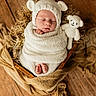 newborn, baby, infant, wrapped, knitted_hat, knitted_toy, teddy_bear, wooden_basket, brown_floor, cozy, sleeping, cute, soft_texture, blanket, portrait, child, indoors, peaceful, warm_colors, resting