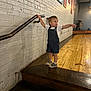 toddler, child, denim_overalls, white_tshirt, pointing, standing, wooden_steps, handrail, brick_wall, indoor, floor, curious, casual_clothing, footwear, short_hair, person, young_child, expression, gesture, lighting