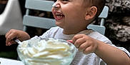 Zayan participe au concours pour gagner de l'argent avec cette photo : toddler, child, smiling, whipped_cream, face, nose, spoon, glass_bowl, table, chair, outdoor, stone_wall, happy, eating, food, person, short_hair, white_shirt, fun, playful