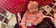 Naya participe au concours pour gagner de l'argent avec cette photo : baby, smiling, infant, headband, pink_clothing, red_pants, socks, blanket, couch, patterned_background, plush, happy, cute, indoors, portrait, child, warm_colors, cozy, sitting, soft_texture