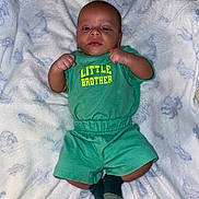 Aemilius is registered to the contest to win money with this photo: baby, infant, child, portrait, lying_down, blanket, green_outfit, shirt, shorts, socks, fists, face, eyes, smiling, indoor, bed, cozy, family, text_on_clothing, photography
