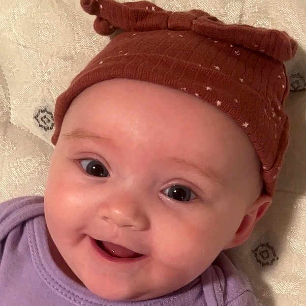 Alyza is registered to the contest to win money with this photo: baby, smiling, face, hat, brown_hat, purple_clothing, infant, cute, child, eyes, happy, portrait, close_up, headwear, baby_girl, soft_fabric, background, skin, cheeks, expression