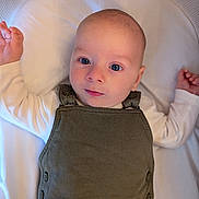 Edward is registered to the contest to win money with this photo: baby, infant, face, eyes, clothing, overall, white_shirt, lying_down, mesh_background, soft_surface, curious, arms_up, cute, child, portrait, indoors, young, person, head, skin