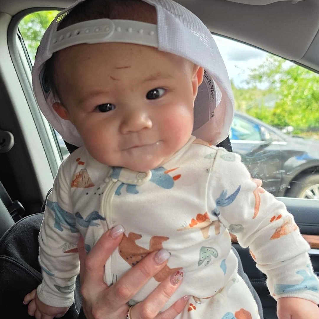 Benji joined the competition — help win amazing prizes! baby, child, dinosaur_onesie, cap, white_cap, car_interior, hand, adult_hand, seat, window, car, trees, greenery, smile, cute, portrait, infant, clothing, person, indoor