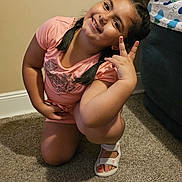 Adrianna is registered to the contest to win money with this photo: child, girl, smiling, peace_sign, braided_hair, pink_shirt, white_sandals, carpet, indoor, living_room, couch, pillows, happy, kneeling, person, casual_clothing, home, portrait, cute, friendly
